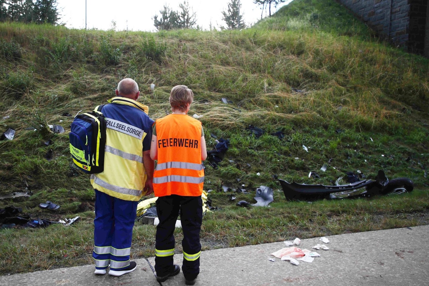 Ein Seelsorger und ein Mitglied der Feuerwehr an einer Unfallstelle auf der Autobahn 9 vor einer Böschung, die mit Trümmerteilen übersät ist.